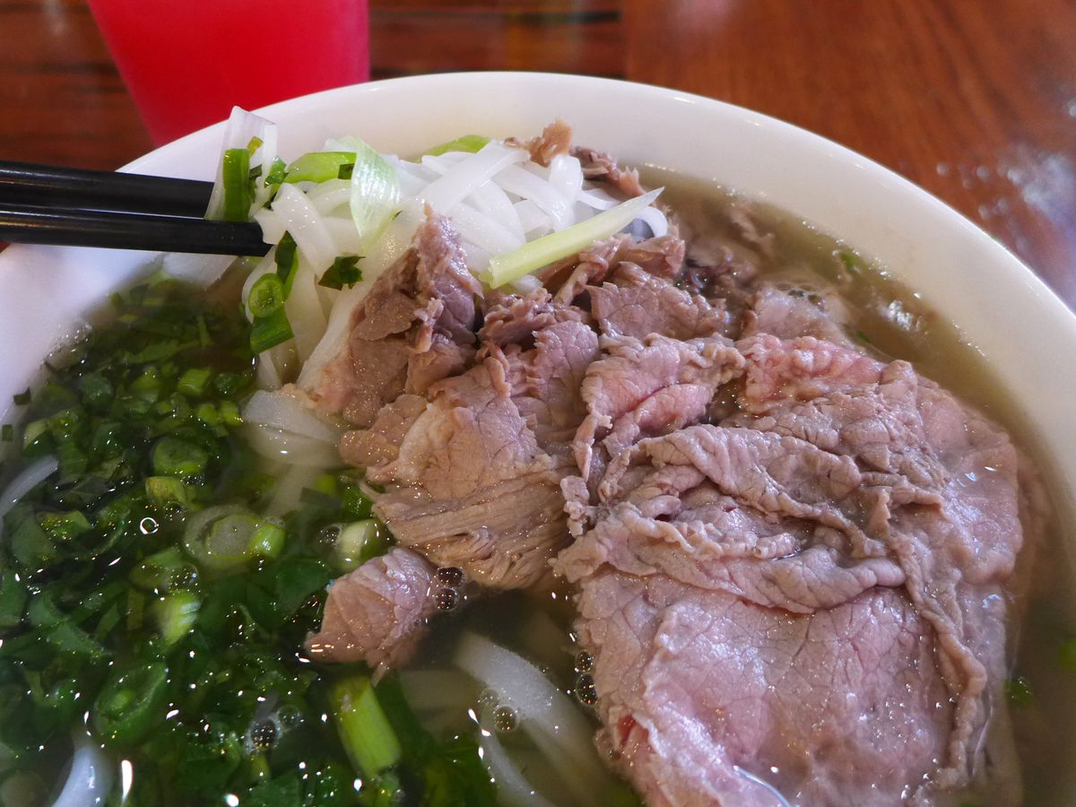 A bowl of soup with herbs and sliced beef pink at the edges, as a pair of chopsticks lifts up some translucent white noodles.