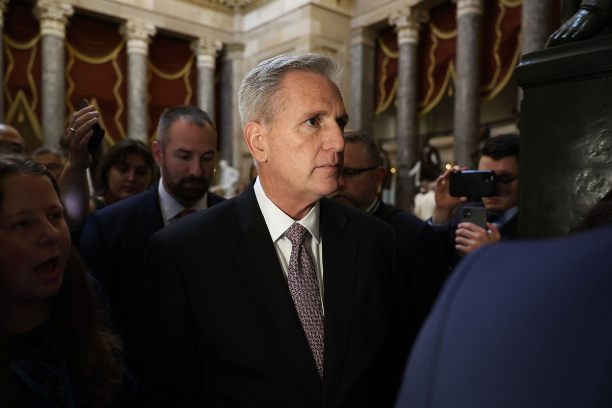 McCarthy, silver haired and clean shaven in black suit, white shirt, and purple tie, wears a neutral expression while surrounded by staff and reporters. The group walk among the Capitol’s columns, from which hang ornate red and gold curtains.