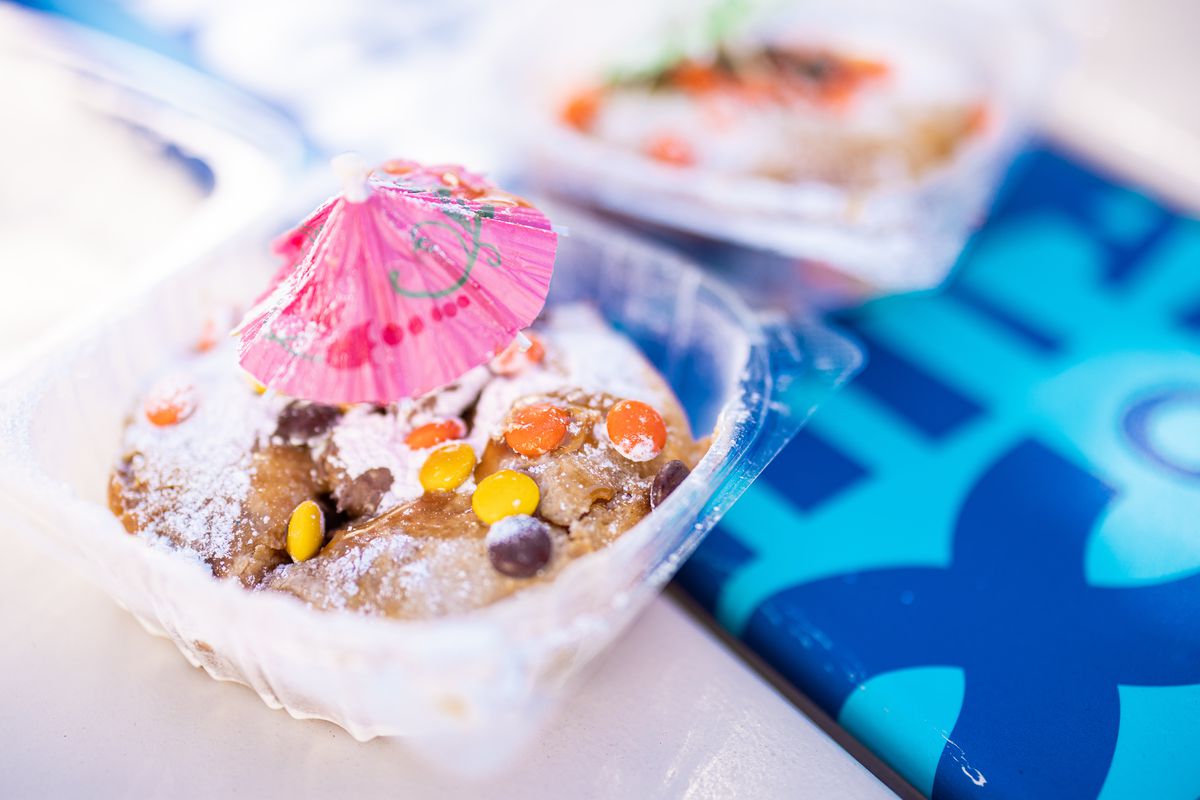 A honey bun that’s decked out with M&amp;Ms, powdered sugar, and a pink paper umbrella sits in a plastic to-go container. 