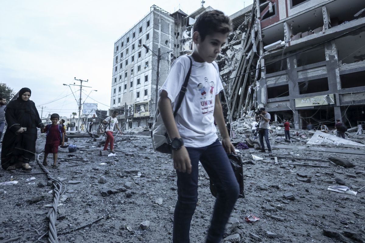 Young children, among others, walk in front of a destroyed, partially collapsed building. The ground is covered in dusty rubble and debris. 