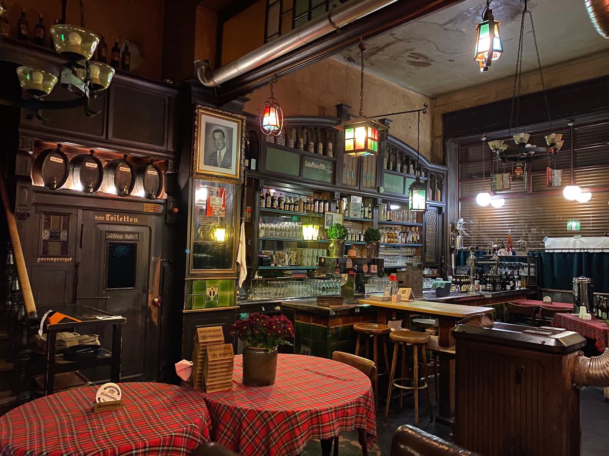 A restaurant interior with high ceilings, wood paneling, small wooden beer kegs lining shelves, a bar, ornate pendant lights, and tables with checkered tablecloths 