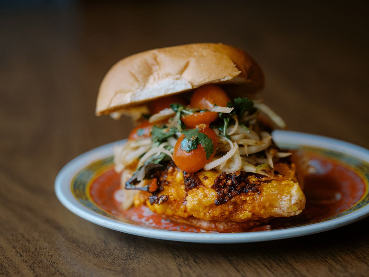 A fried chicken sandwich topped with tomatoes and papaya slaw on a red and white plate.