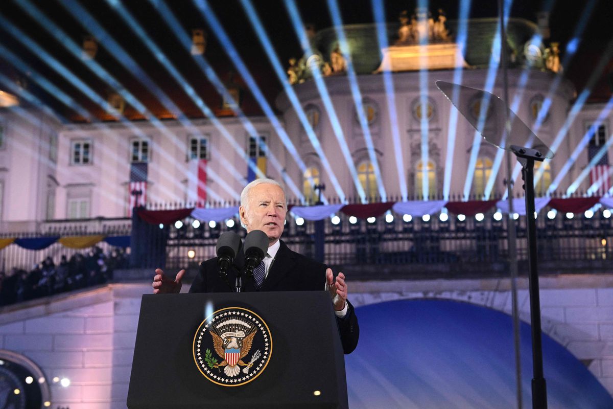 President Biden speaks outdoors at a podium with the US presidential seal on it, wearing a dark suit and white shirt. Behind him, lights display a pattern of blue stripes across Warsaw’s Royal Castle museum. 
