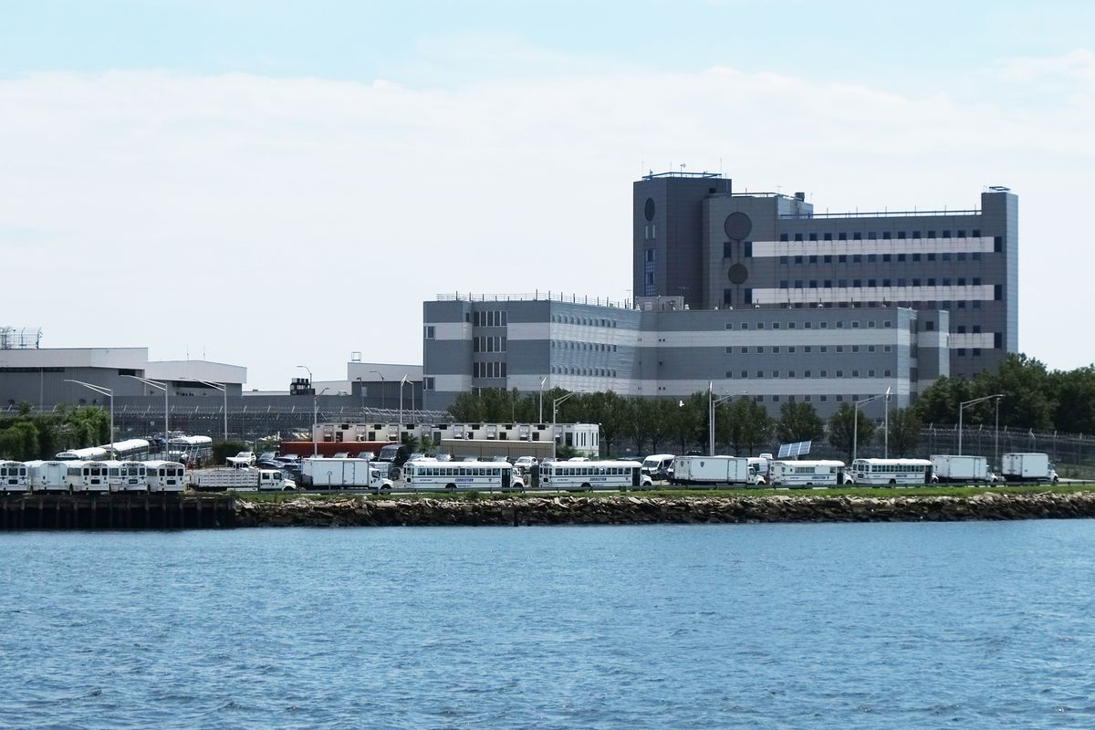 A high-rise jail on Rikers Island with white Department of Correction buses lined in front of it.