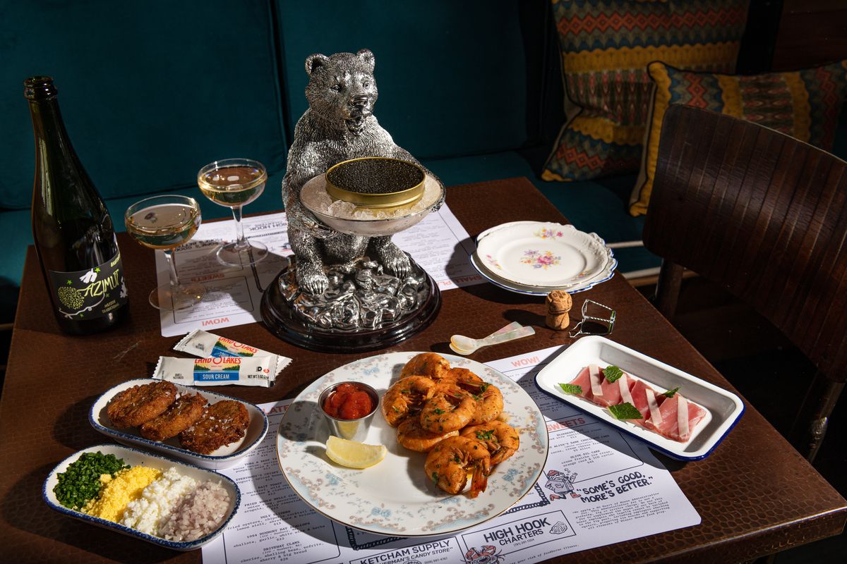 A crudo, shrimp, and a few other seafood dishes sit on a dark wooden table on a paper placement. A bear figurine holds a caviar tin.