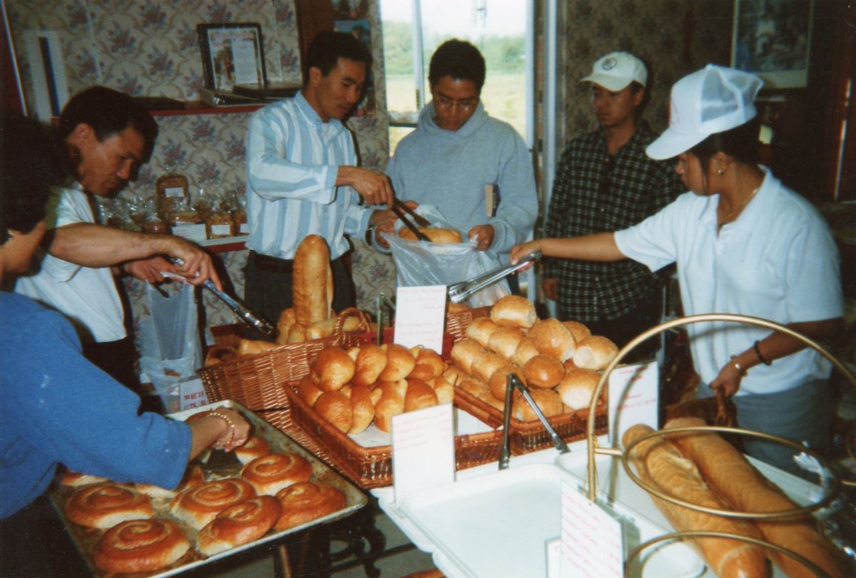 A group of people standing around a bakery counter selecting rolls of bread, baguettes, and swirled pastries with metal tongs.