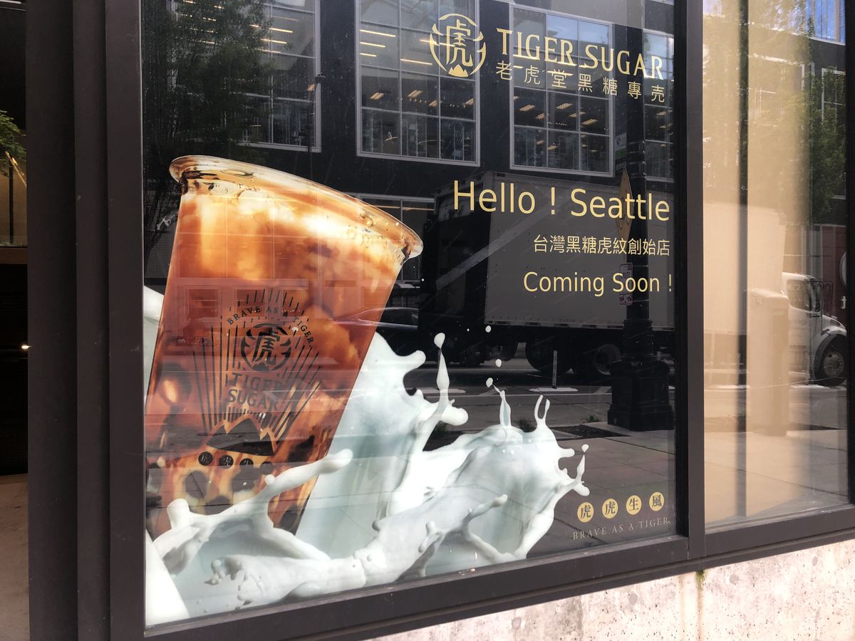 A glass storefront with a big picture of a cup of bubble tea streaked with black syrup, with milk splashing around the cup and the words “Hello! Seattle, Coming Soon,” written next to the image.