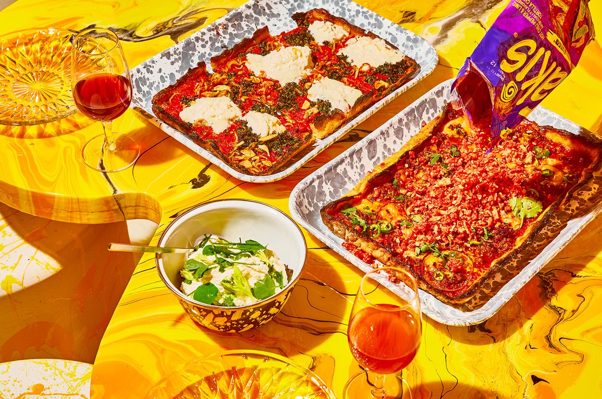 Two pan pizzas on a bright-yellow table alongside wine glasses; an unseen hand pours Takis from a bag onto one of the pizzas.