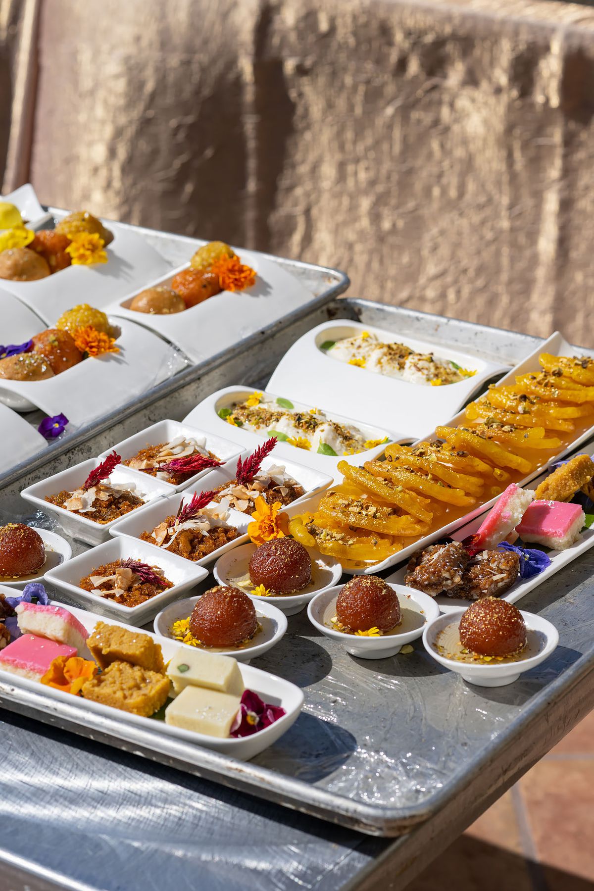 A silver counter covered with small plates in different shapes and sizes, each featuring traditional and non-traditional Indian celebration dishes.