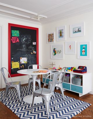 dětský pokoj s modrobílým kobercem chevron, čtyři hliníkové židle Tolix, bílý tulipánový stůl, tabule s červeným rámečkem, ilustrované kresby na zdi, a lavička složená z kójí
