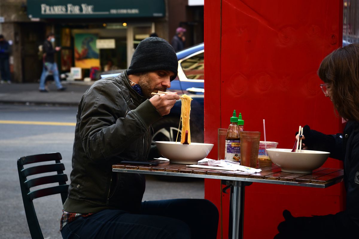 A man eats ramen at an outdoor restaurant table wearing a winter hat and jacket.