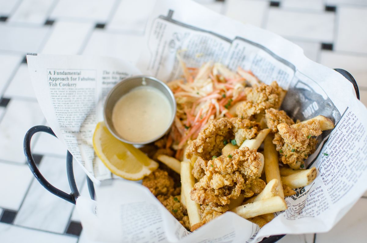Fried clams, fries, a lemon wedge, and a silver cup of remoulade sit on newspaper in a black basket. A white and black tiled floor is visible underneath.