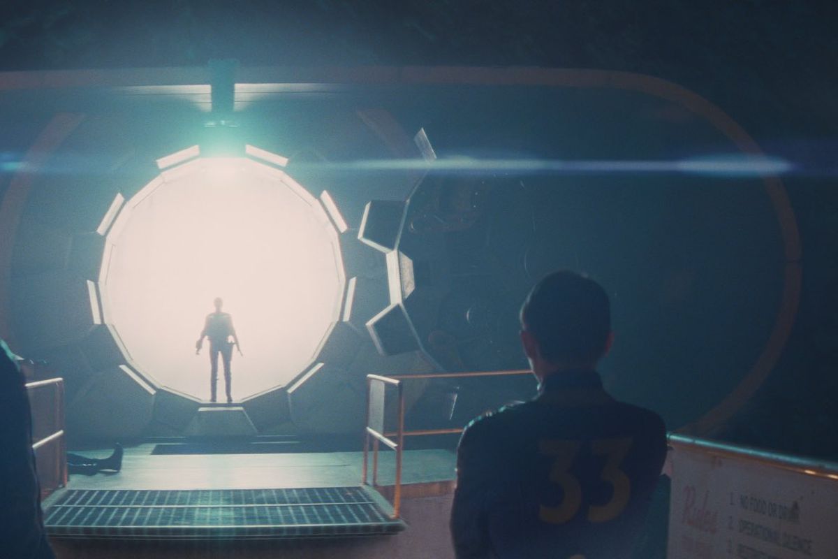A silhouette stands in front of the blinding white light flooding into an underground shelter from the open vault door of Vault 33 while three people in denim jumpsuits with 33 on the back look on.