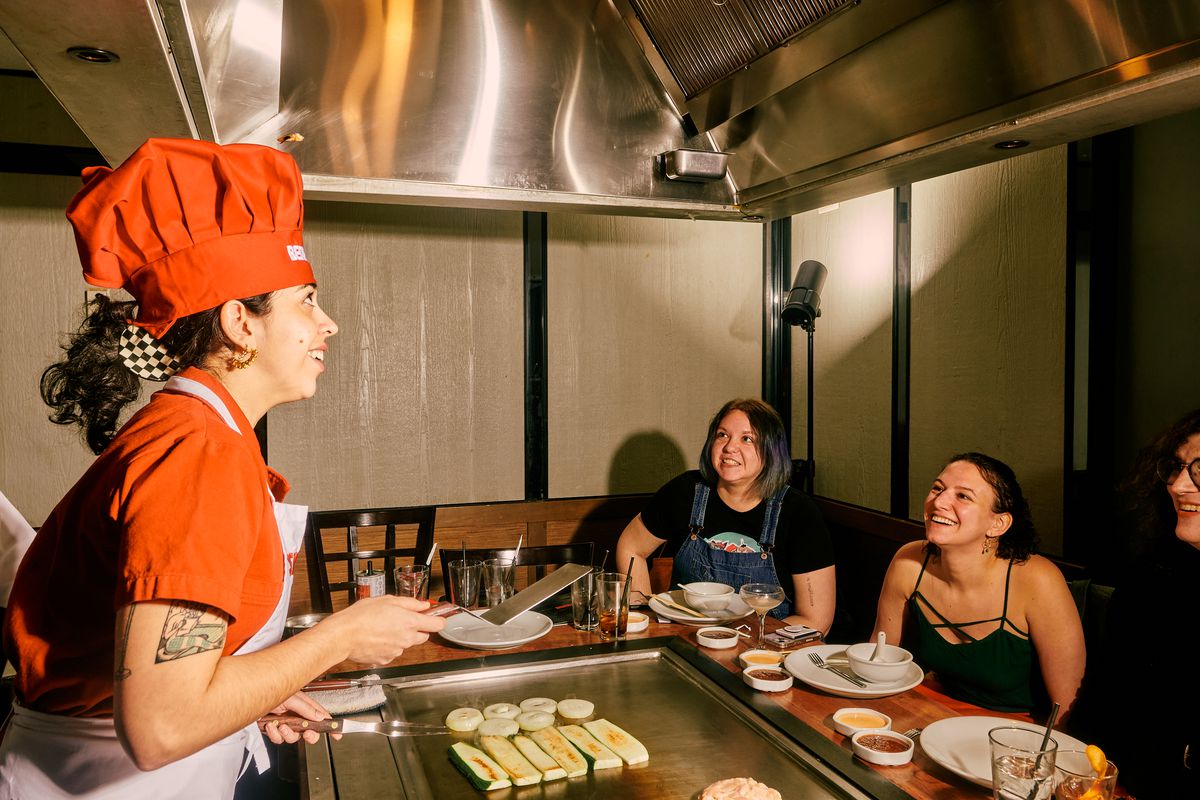 Jaya, standing behind Benihana’s grill, flips a shrimp tail into her chef’s hat as her friends look on, smiling.