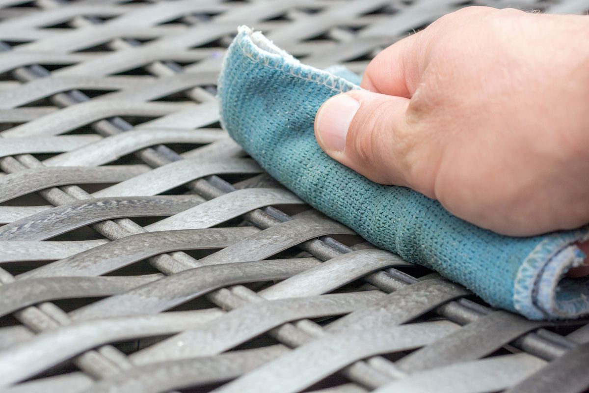 A hand using a blue cloth to wipe down a piece of vinyl wicker furniture. 