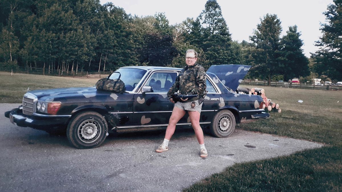 Richard Davis, a white man in shorts and a camo jacket, stands in front of a camo-painted car parked on a driveway next to a large lawn.