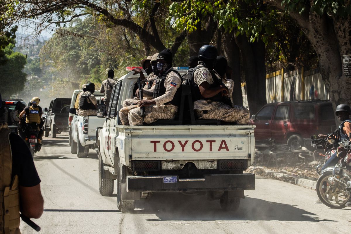 Armed police officers sit in the back of a pick-up truck, driving along a dusty road.