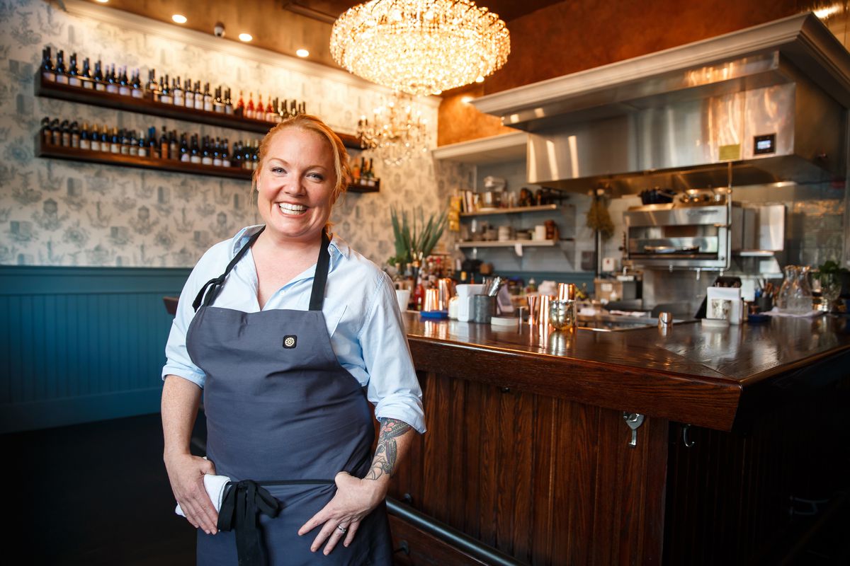 A woman wearing a blue apron over a blue button-down shirt smiles inside a restaurant with a chandelier, dark wood, and blue accents.
