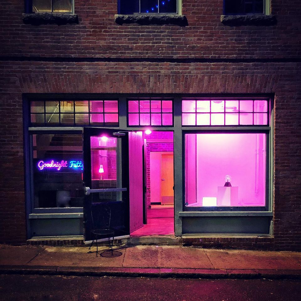 Exterior of a storefront in a brick building at night. The door is open and the space is empty but glows neon pink. A neon sign that says “Goodnight Fatty” in cursive is visible in one window.
