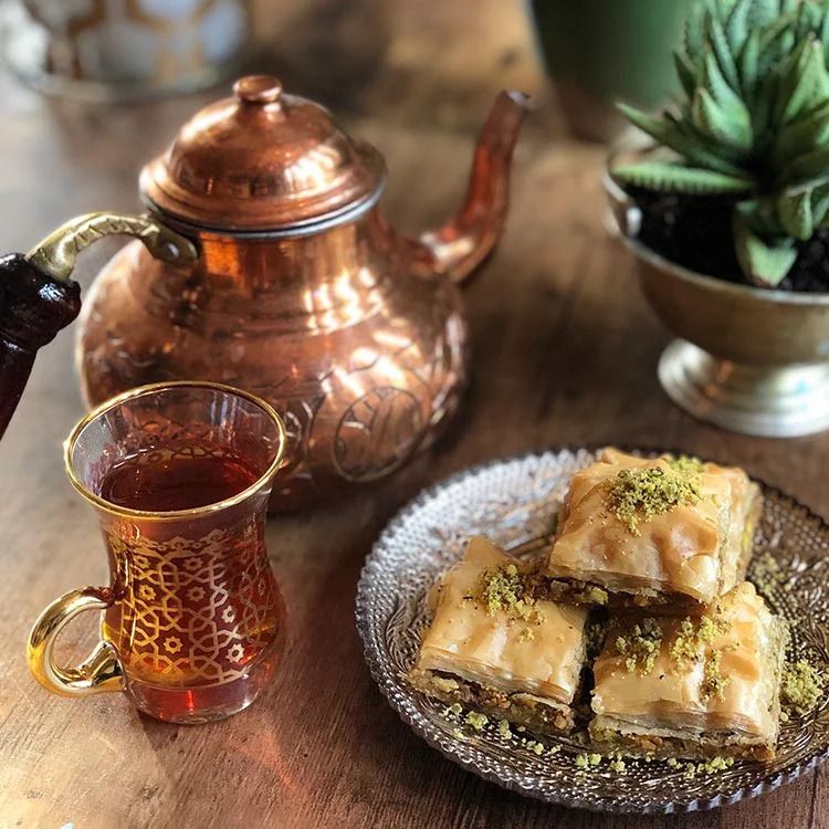 A stack of pieces of baklava on a decorative glass plate, beside an ornate glass mug of tea and a copper teapot