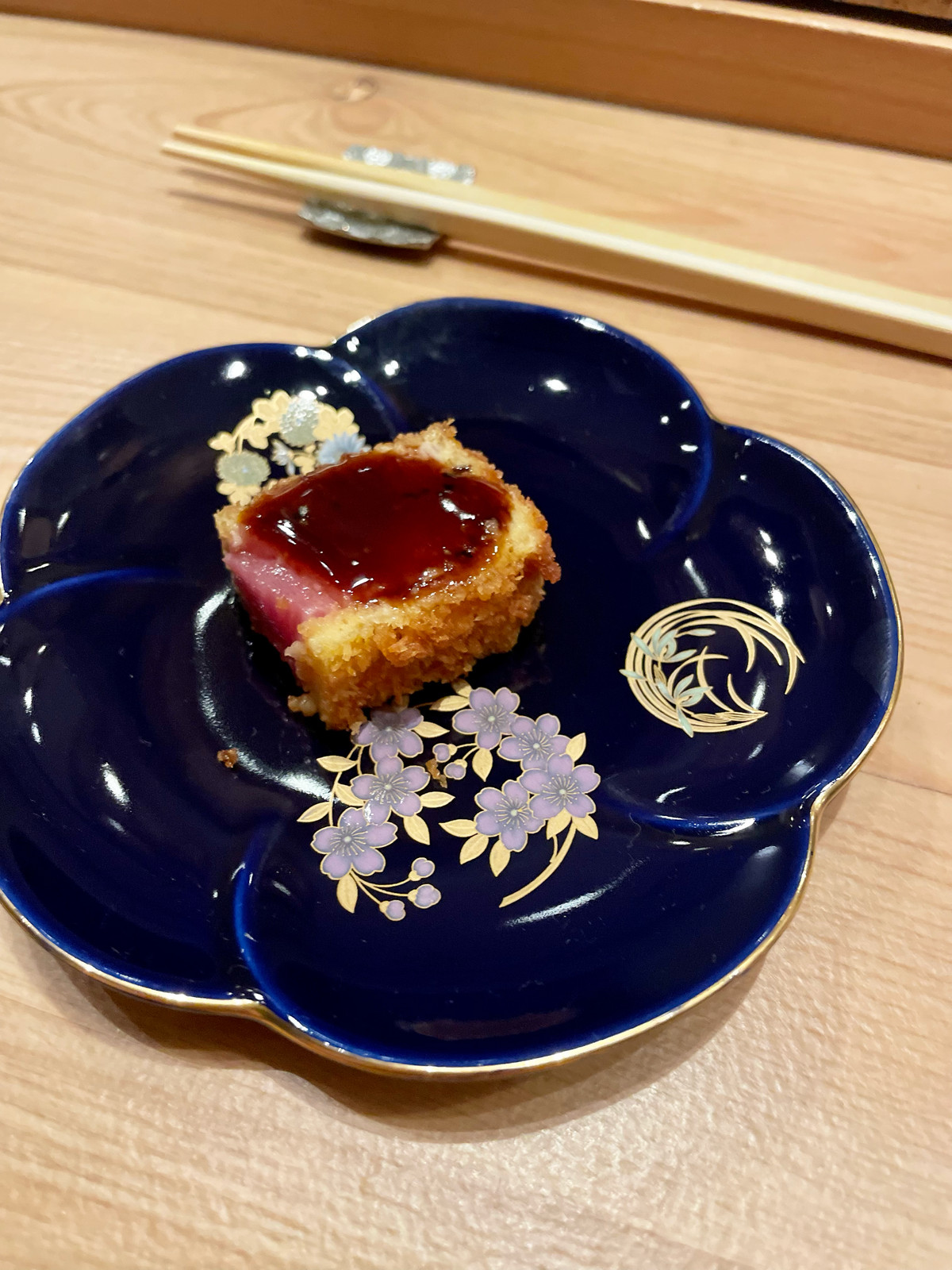 On a navy plate with gold and purple flowers, a piece of toro that has been crusted in Panko and fried, then topped with a sauce, sits.
