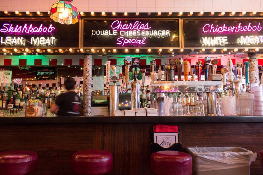 A restaurant interior features red leather stools at a bar. Neon signage over a mirror in the background advertises Charlie’s double cheeseburger special.
