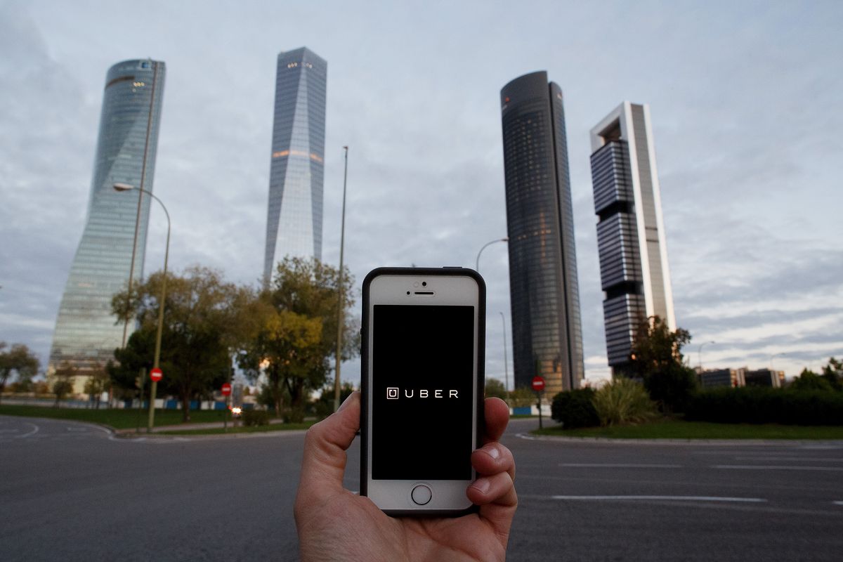 A hand holds a mobile phone showing Uber’s app screen, with Madrid, Spain, buildings in the background. 