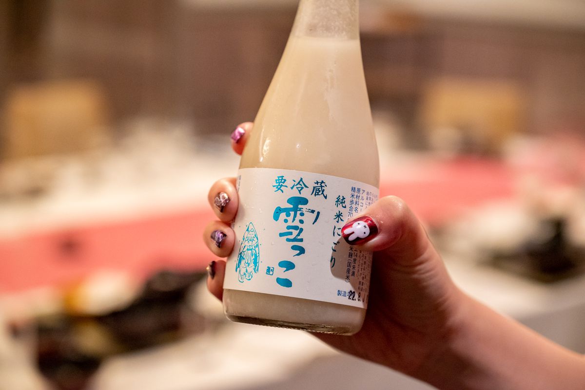 A hand with painted nails holds up a cloudy bottle of sake.