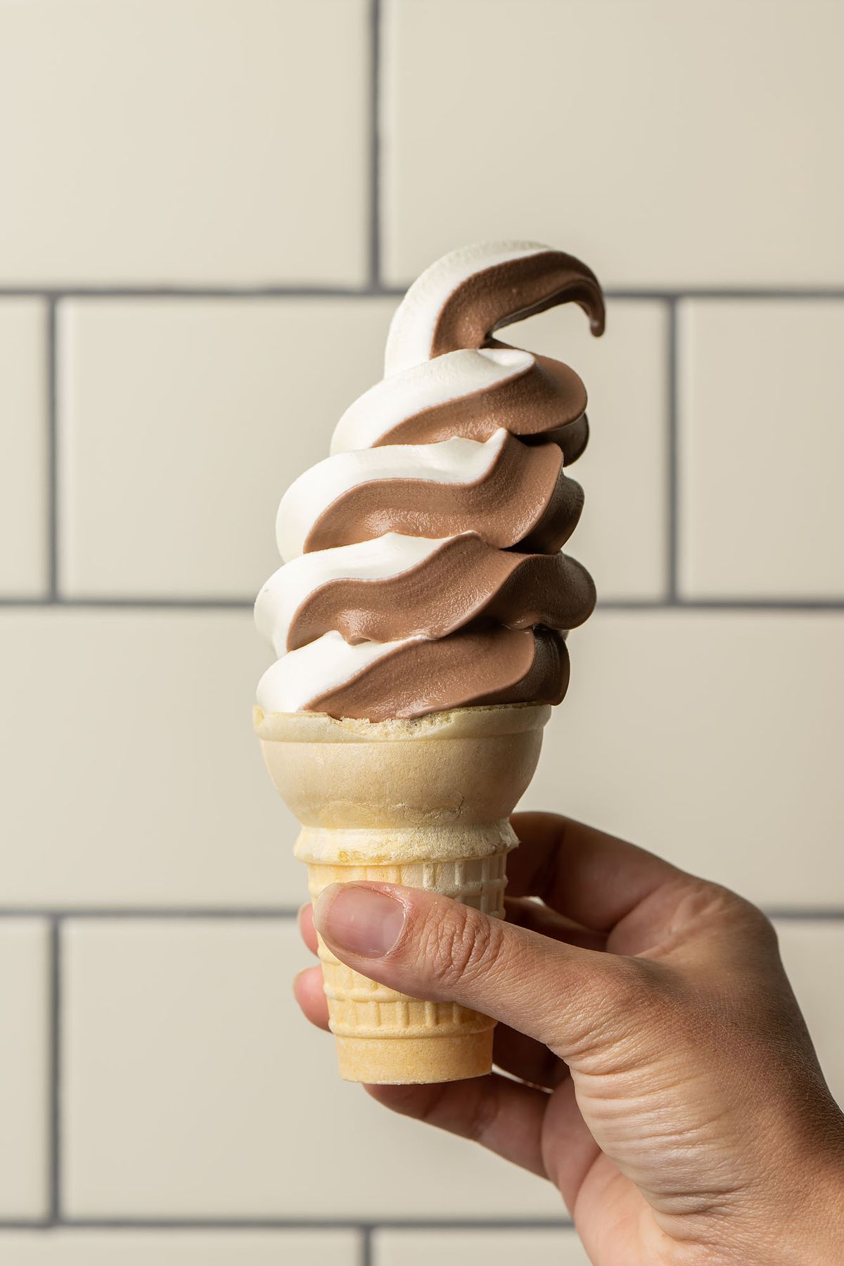 A hand holds up swirled soft serve against a tile background.