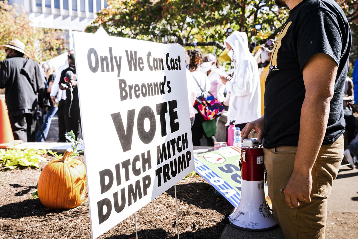 The photo focuses on a white sign with bold black text that been stuck into a patch of dirt. A pumpkin is behind it. The sign reads, “Only We Can Cast Breonna’s VOTE DITCH MITCH DUMP TRUMP.” A Black man’s torso can be seen next to the sign. He has a bullhorn in one hand, and is wearing a black t-shirt. The sun is shining brightly, and in the background is a crowd of people, all of them in black or white. 