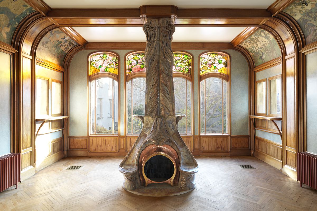 Ceramic stove in middle of room with stained glass windows. 