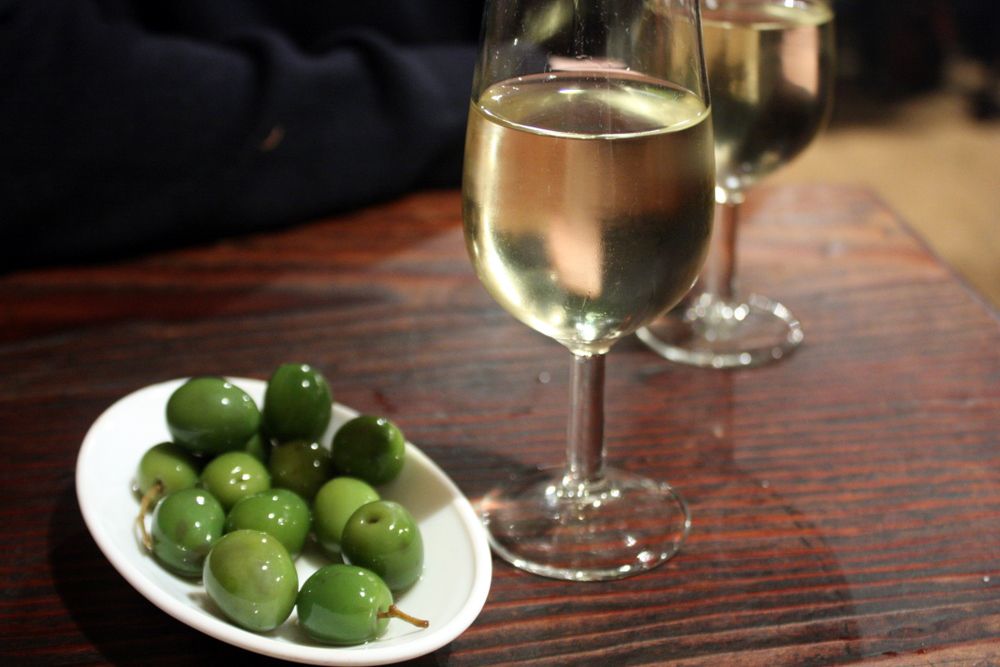 A glass of sherry sits next to a small plate of olives on a wood bar with someone leaning nearby.