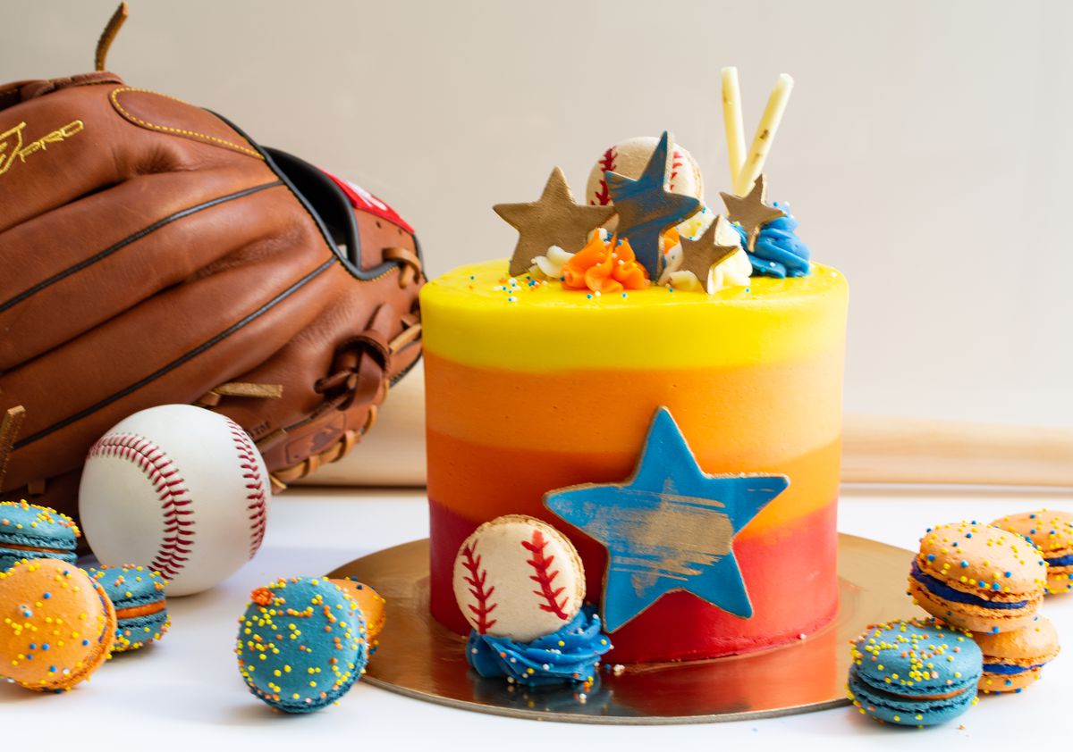 An Astros-themed 6-inch cake decorated with stars and a baseball, with blue and orange macaroons with sprinkles nearby.