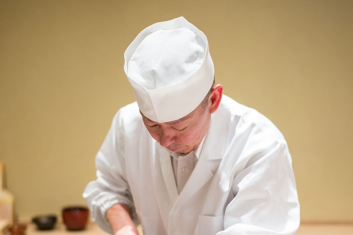 Eiji Ichimura wearing chef’s whites and a white hat behind a sushi counter.