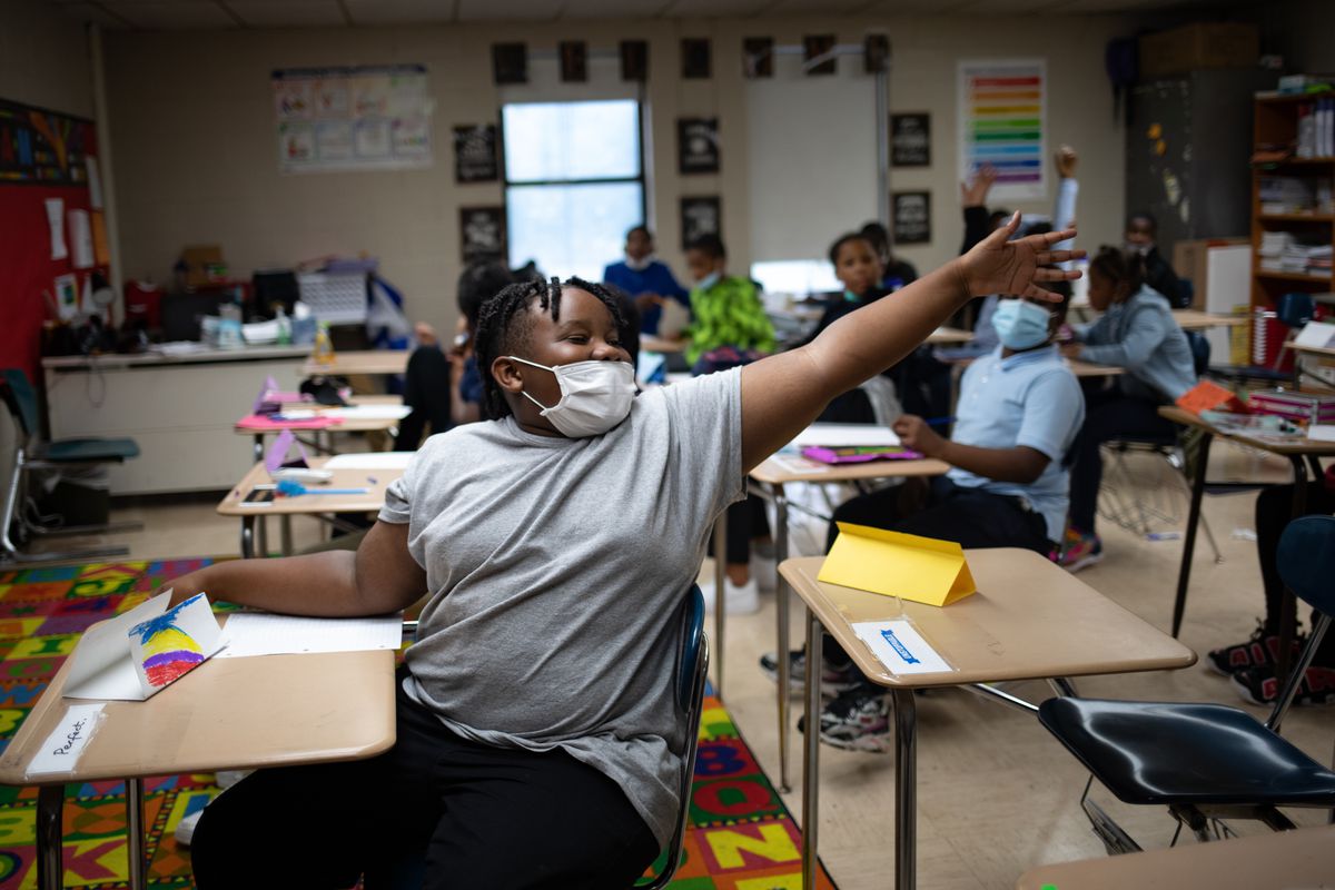 A student emphatically raises their hand during an exercise in class. The student and their peers are all wearing protective masks at their desks in the classroom.