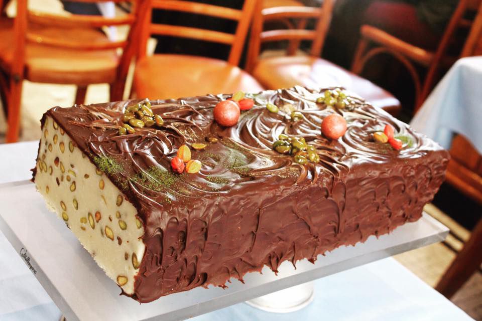 A large slab of nougat, dotted with toppings, sits on a cake stand with one end slice missing.