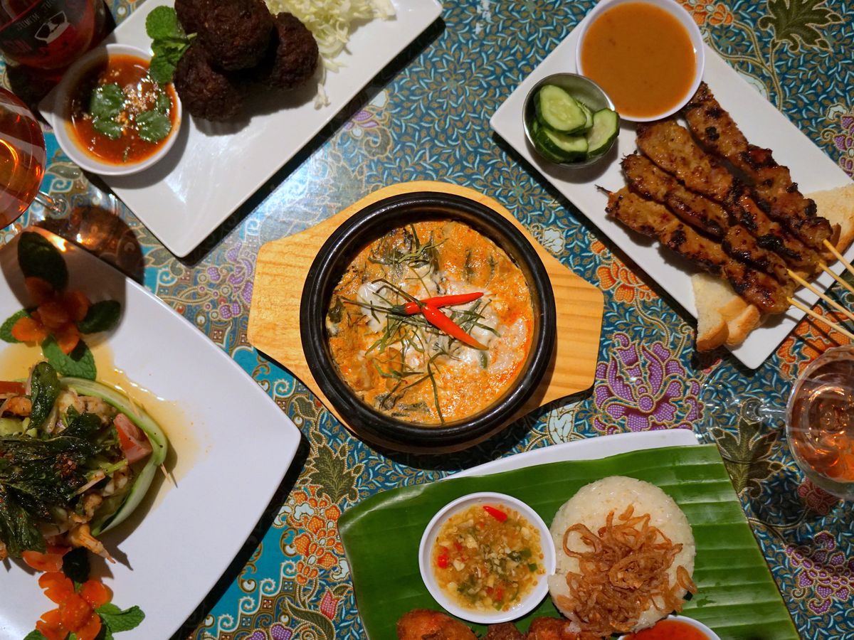 A floral table-top covered with an assortment of Thai street food dishes, including a white plate with meat skewers, peanut sauce, and cucumbers, a black stone bowl filled with curry and sliced chiles, and a banana leaf topped with rice and chile-laden fish sauce.