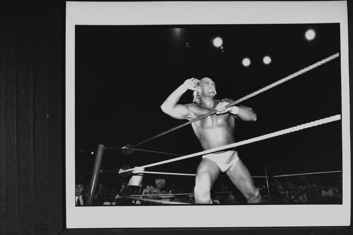 Une photo en noir et blanc montre Hulk Hogan dans un ring de catch à son apogée, appelant la foule à faire du bruit.