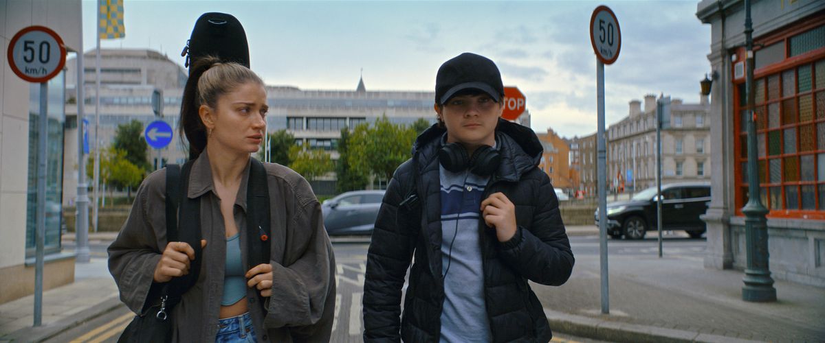Flora, played by Eve Hewson, walks through Dublin in a jacket with a guitar case strapped to her back next to her son, Max, in the Apple TV Plus film Flora and Son