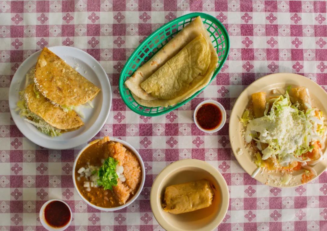 A checkered tablecloth set with Mexican dishes including tortillas, flautas, and tacos.