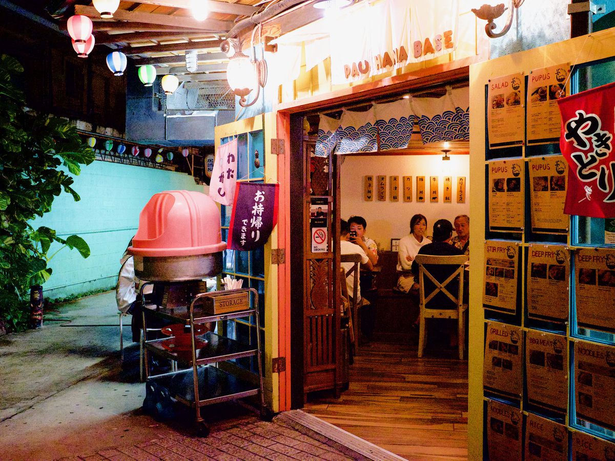 A restaurant entrance, decorated with string lights and Japanese signs, with a turquoise alleyway leading away to one side
