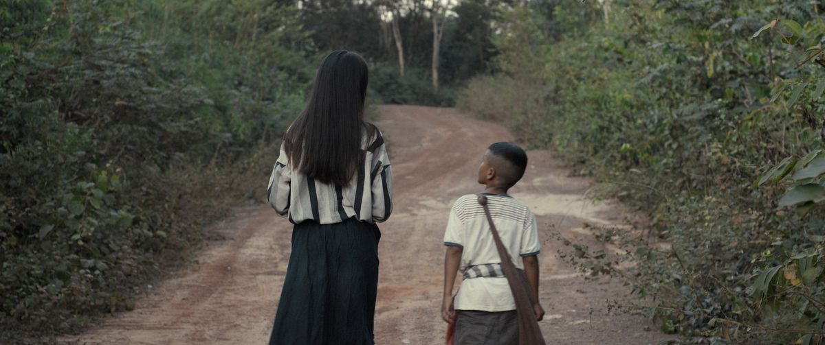 A little Laotian boy and his spirit companion are walking along a dirt road through the forest in Mattie Dos The Long Walk