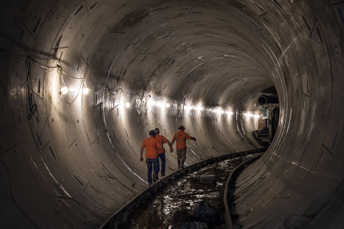 Workers in hardhats and vests walking through a dark subway tunnel with flashlights.
