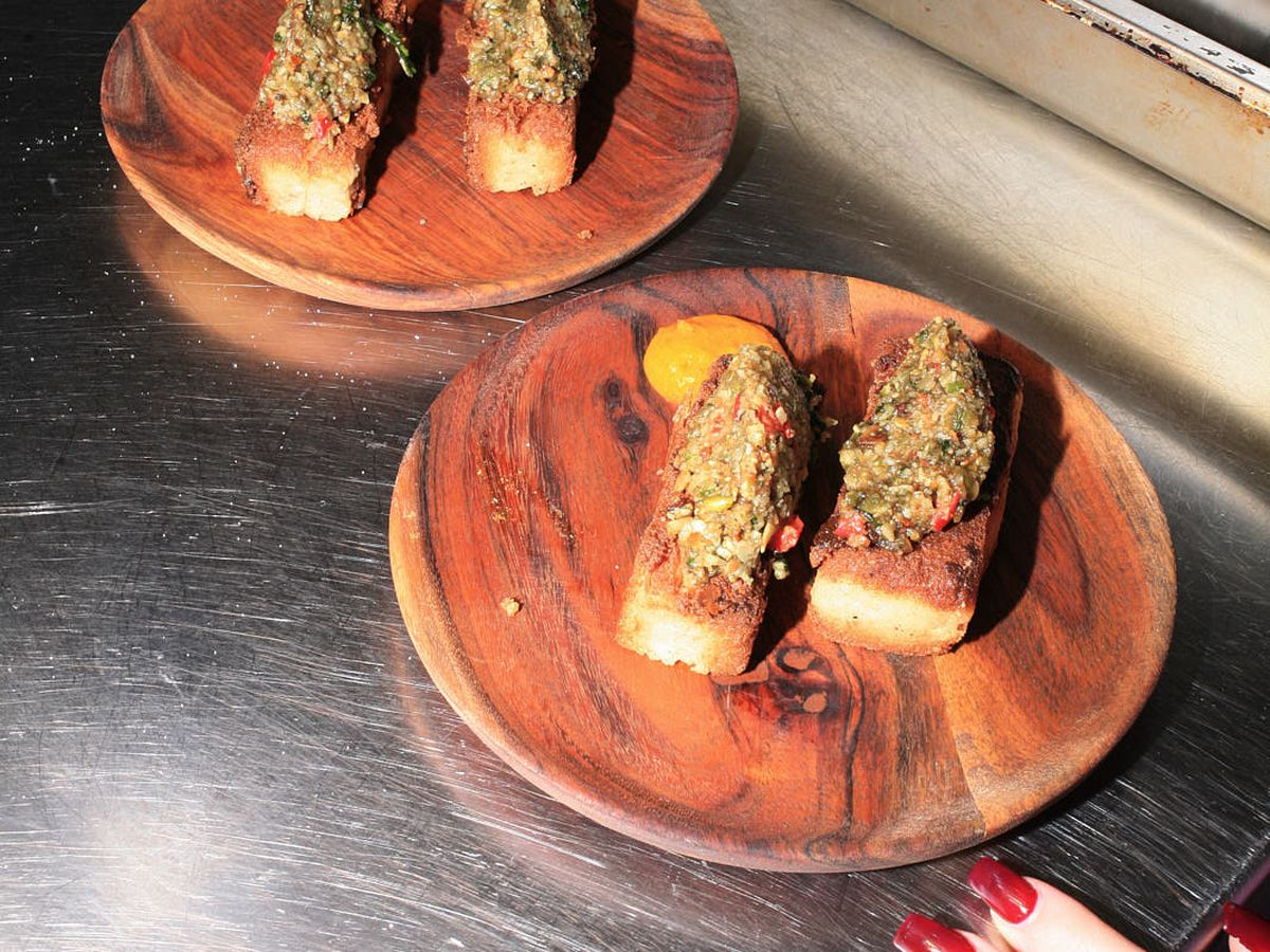 Two wooden plates with two portions each of ekuru with pumpkin seed pesto and scotch bonnet sauce, the sauce vivid orange and to the side of the slice of ekuru, and the pesto, dark green, on top.