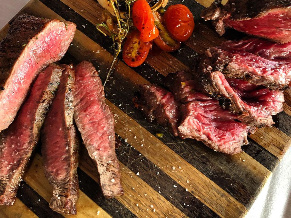 Two different local cuts of beef on a wood board with roasted cherry tomatoes and garlic.