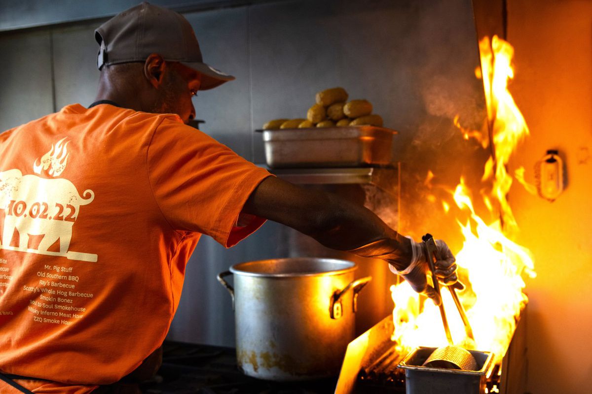 A man in an orange shirt, baseball cap and apron reaching tongs into a flaming pan.