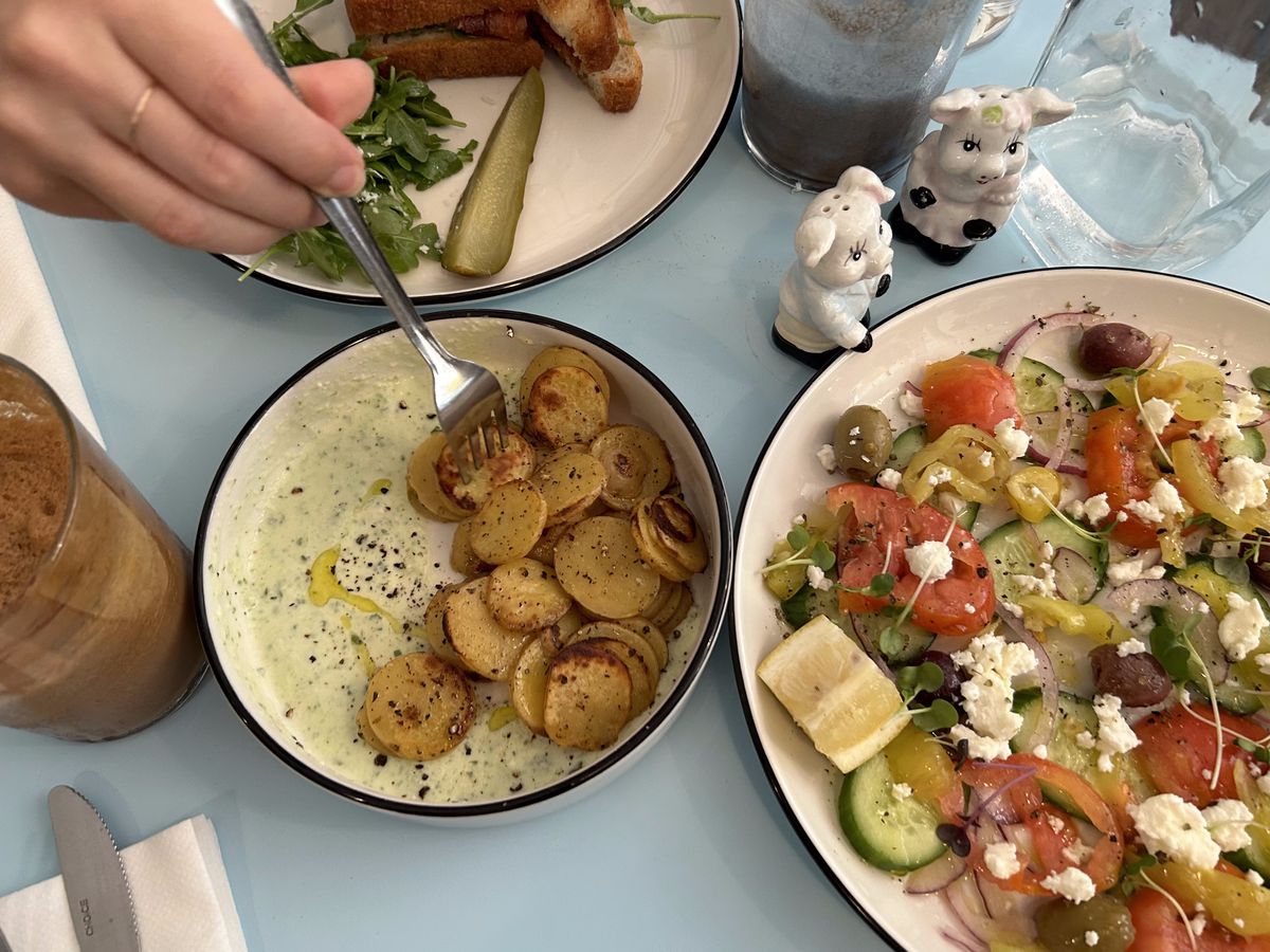 A hand holding a fork impales a small sliver of potato next to a colorful salad with tomato, feta, and other ingredients.
