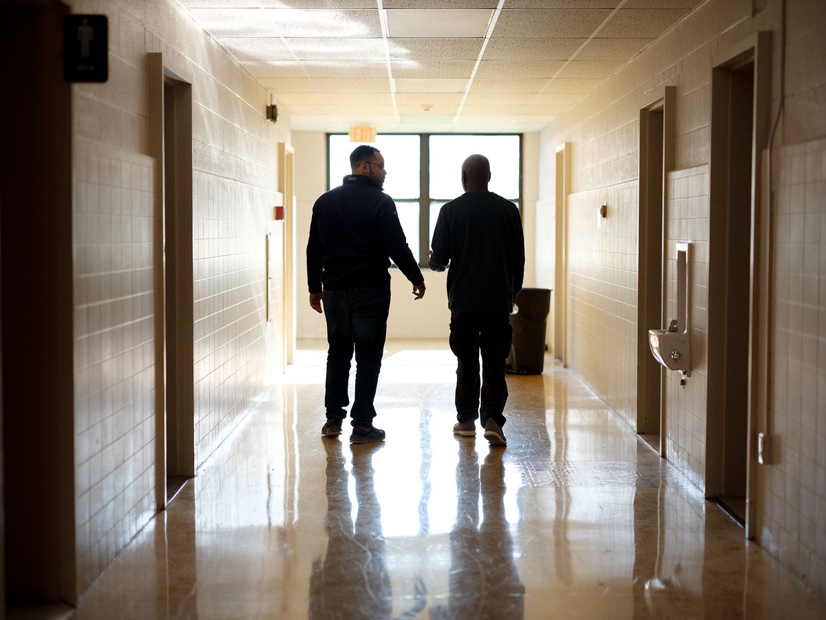 A pair of Black men talking with each other walk down a tiled hallway, seen from behind.