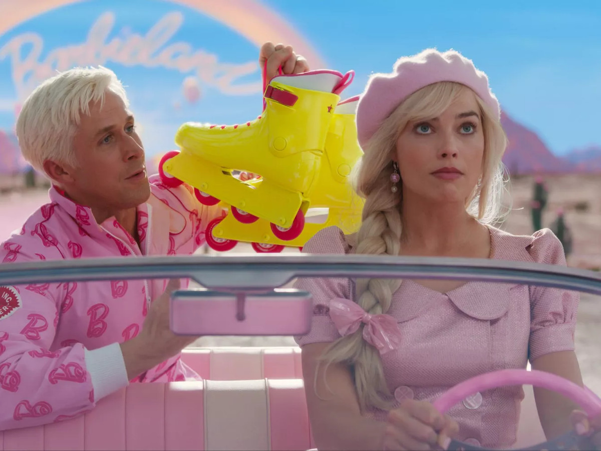 The two actors are in a car, driving away, “Barbieland” faintly seen in the distance. Ken holds up yellow rollerblades.