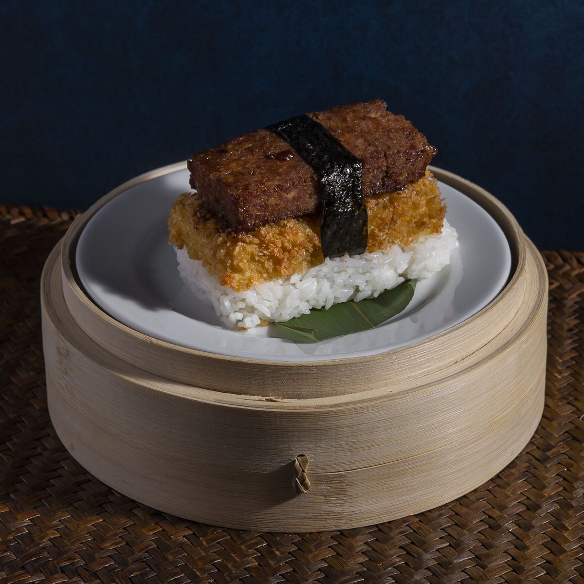 A rectangle of rice is topped with egg and spam, wrapped in a thin slice of seaweed, and presented atop a steamer basket.